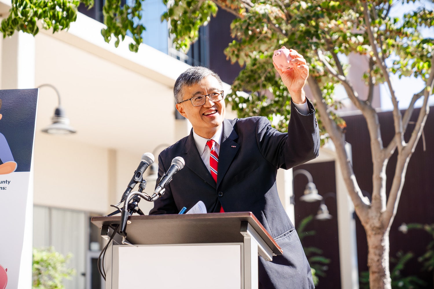 Photo of Supervisor Otto Lee speaking during MedAssist press conference, smiling, holding a stress-ball piggy bank.